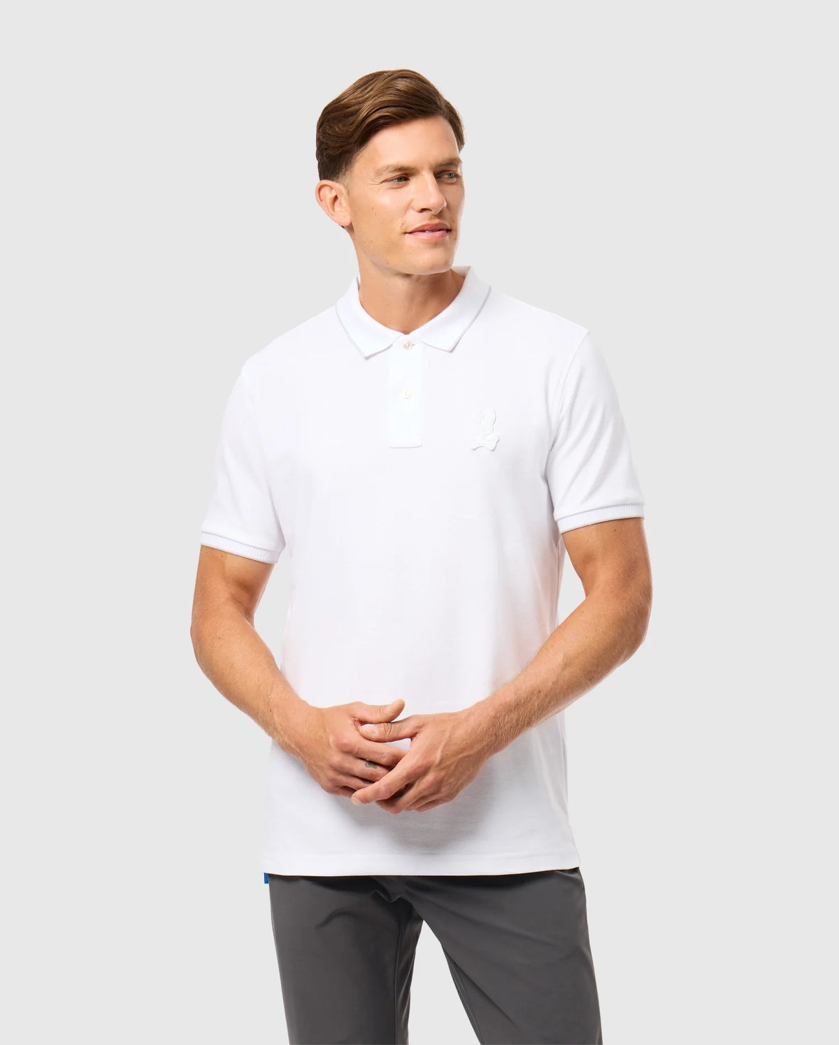 A man with short brown hair wears a white Psycho Bunny Men's Kingston Pique Polo Shirt (B6K764D200) made of Pima cotton, paired with gray pants, while standing against a neutral gray background. His hands are clasped together in front of him, and he is looking off to the side with a slight smile on his face.