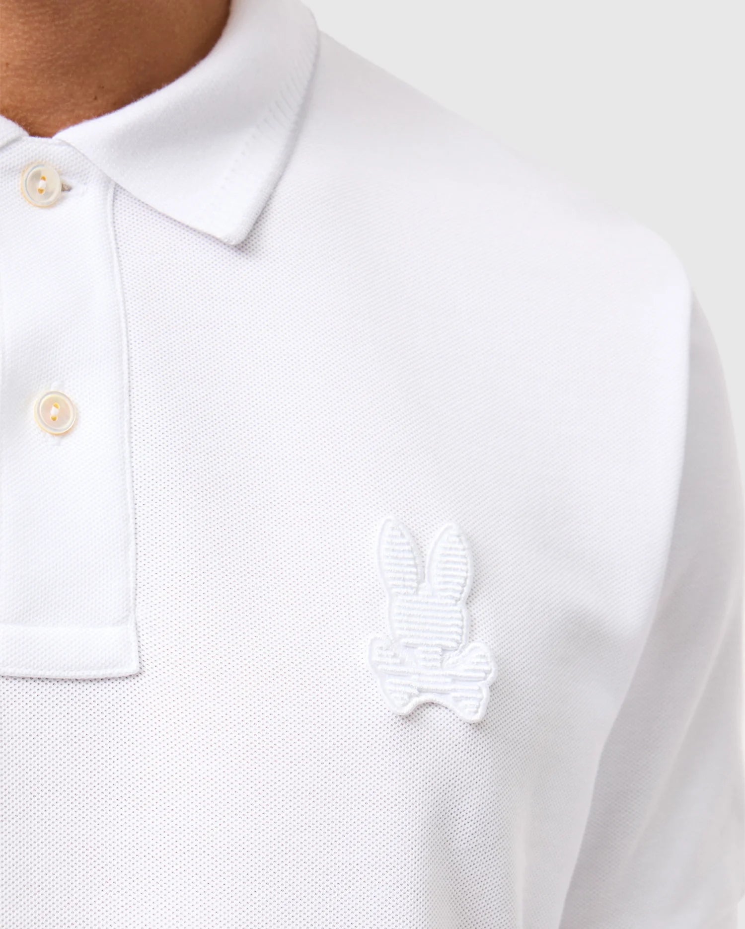 Close-up of a person wearing a white Psycho Bunny MENS KINGSTON PIQUE POLO SHIRT - B6K764D200, made from Pima cotton, featuring button details and an embroidered Bunny patch with crossed bones on the left side of the chest. The background is a neutral gray.