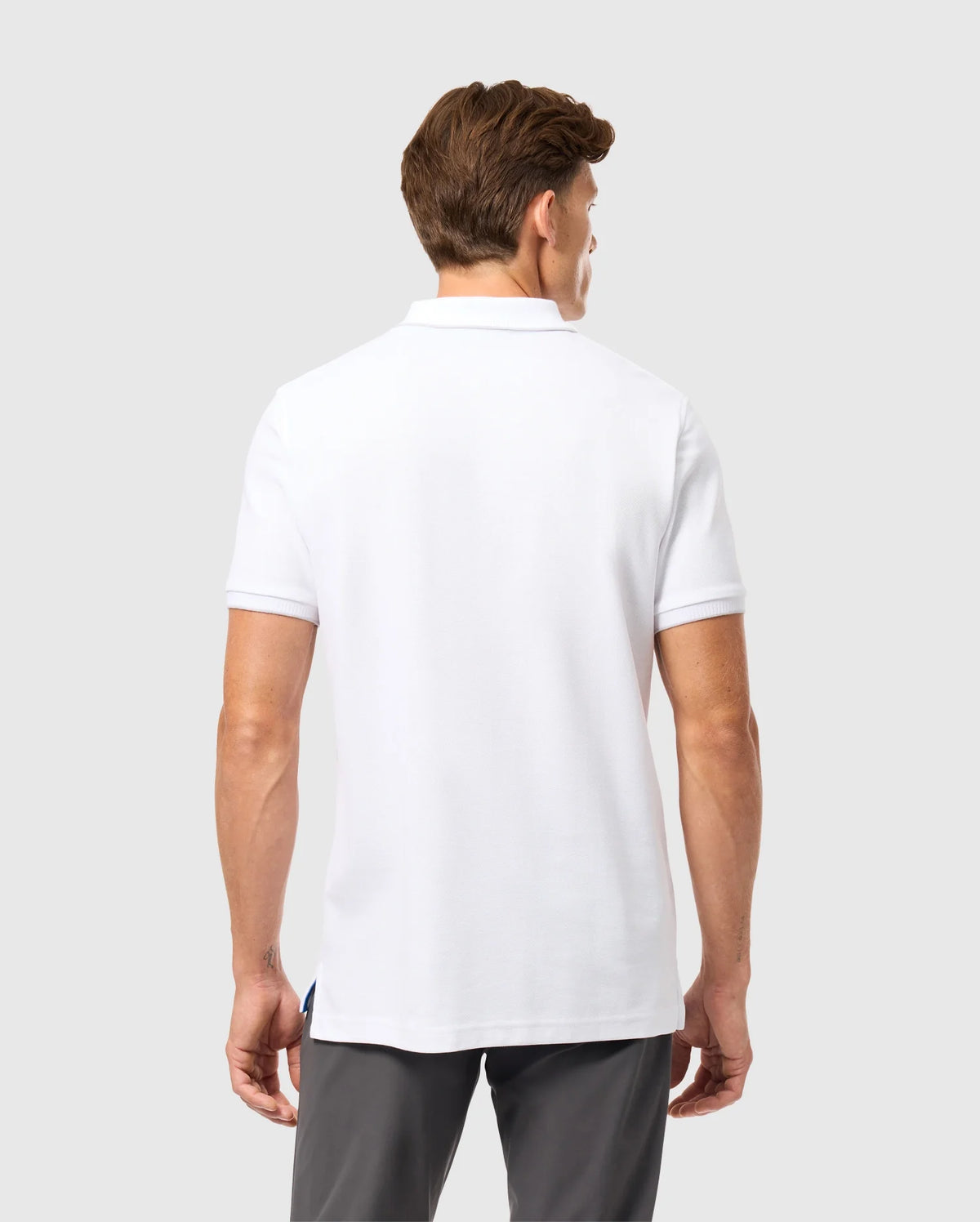 A man with short brown hair is facing away from the camera, wearing a white Psycho Bunny Men's Kingston Pique Polo Shirt (B6K764D200) with an embroidered Bunny patch, along with grey pants. The background is plain and light-colored.