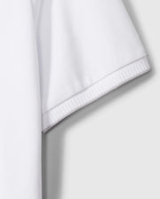 A close-up of the sleeve on the MENS KINGSTON PIQUE POLO SHIRT - B6K764D200 by Psycho Bunny highlights the textured Pima cotton fabric and detailed hem stitching. The white, short-sleeved polo shirt is crafted from light, breathable material. An embroidered Bunny patch adds a touch of charm against the flat, neutral gray background.