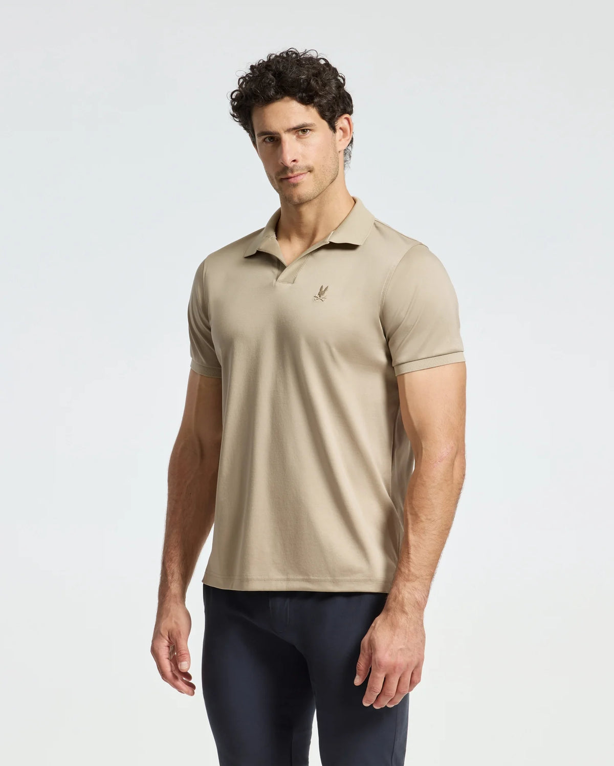 A man wearing a Psycho Bunny MENS BROADWAY PIMA INTERLOCK JOHNNY COLLAR POLO - B6K891D200 in beige stands against a plain light background. His hair is curly, and he looks directly at the camera, pairing his outfit with dark pants.