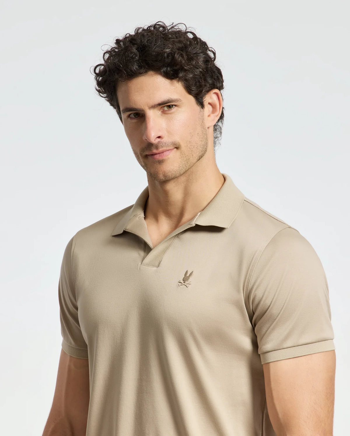 A man with curly dark hair is wearing the Psycho Bunny MENS BROADWAY PIMA INTERLOCK JOHNNY COLLAR POLO in beige, crafted from soft pima cotton and featuring a small embroidered logo on the chest. He stands against a plain light background, looking slightly to the side with a neutral expression.