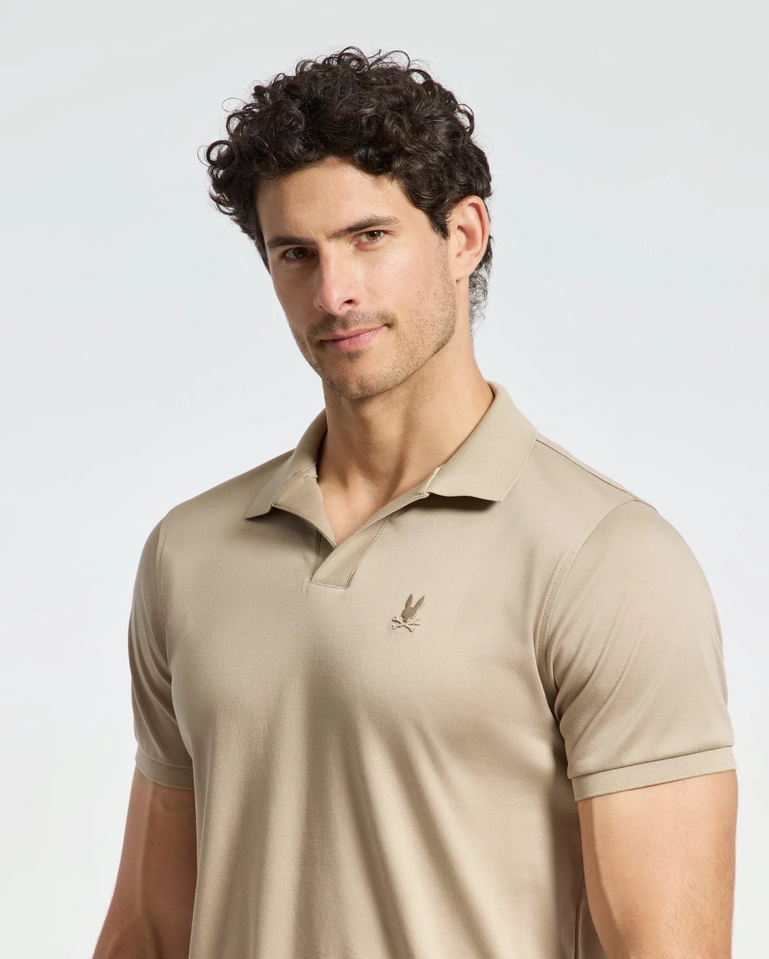 A man with curly dark hair is wearing the Psycho Bunny MENS BROADWAY PIMA INTERLOCK JOHNNY COLLAR POLO in beige, crafted from soft pima cotton and featuring a small embroidered logo on the chest. He stands against a plain light background, looking slightly to the side with a neutral expression.