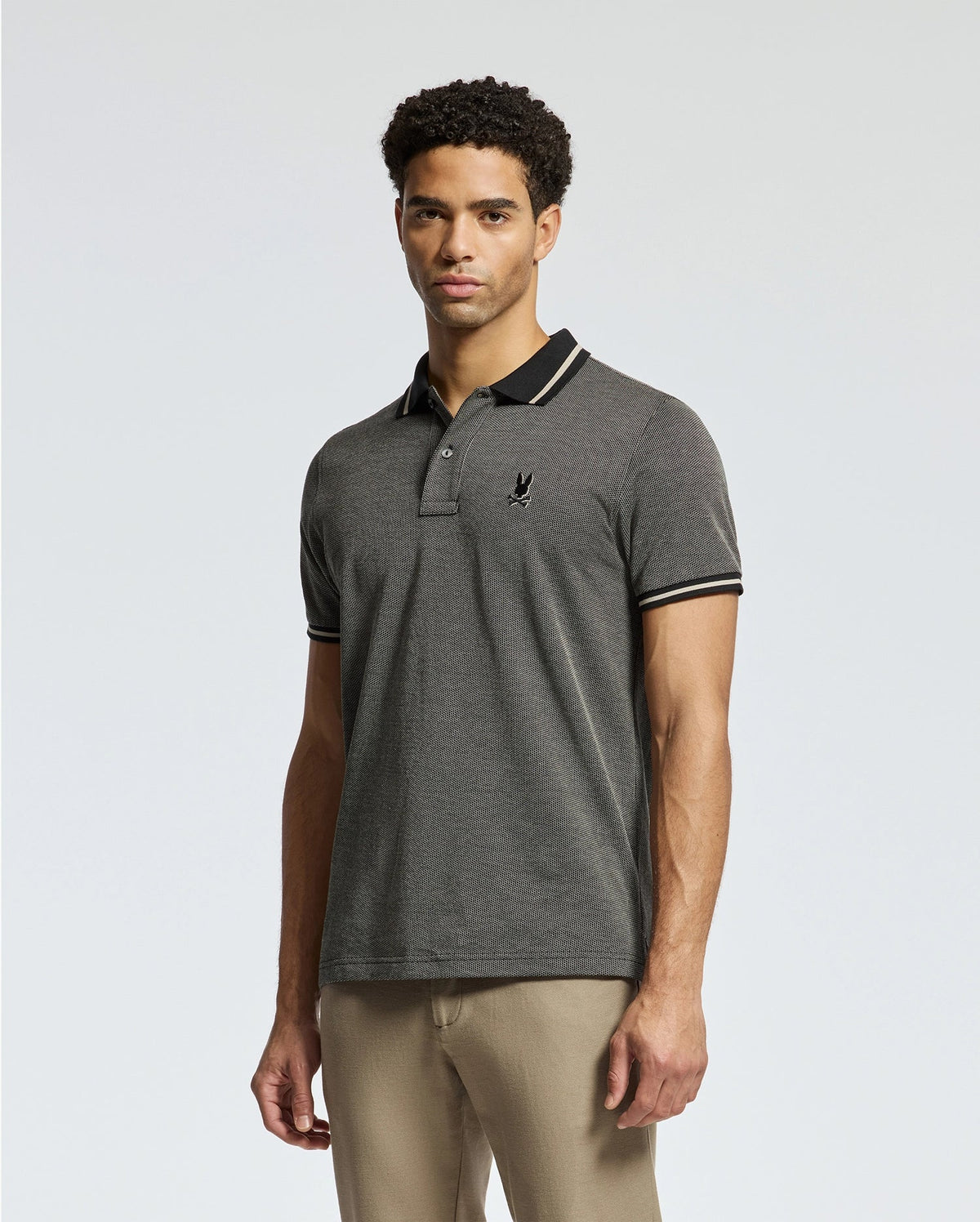 A man stands against a light gray background wearing the Psycho Bunny Men's Warsaw Jacquard Pique Polo (B6K901E200) in charcoal gray, made from luxurious Peruvian Pima cotton. The polo features black trim and an embroidered logo on the chest, which he pairs effortlessly with beige pants.