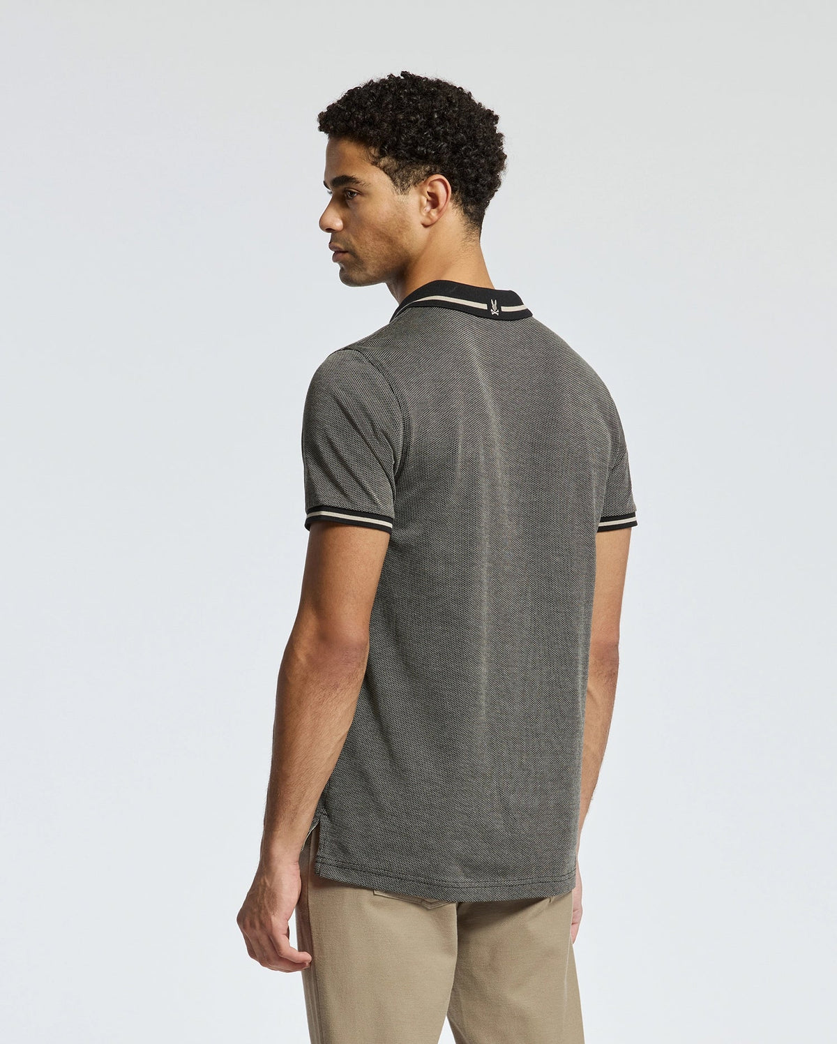 A person poses sideways wearing the Psycho Bunny MENS WARSAW JACQUARD PIQUE POLO - B6K901E200, highlighting the black and white trim on the collar and sleeves of their gray shirt. Their short curly hair complements the look, which is paired with beige pants against a plain light-colored background.