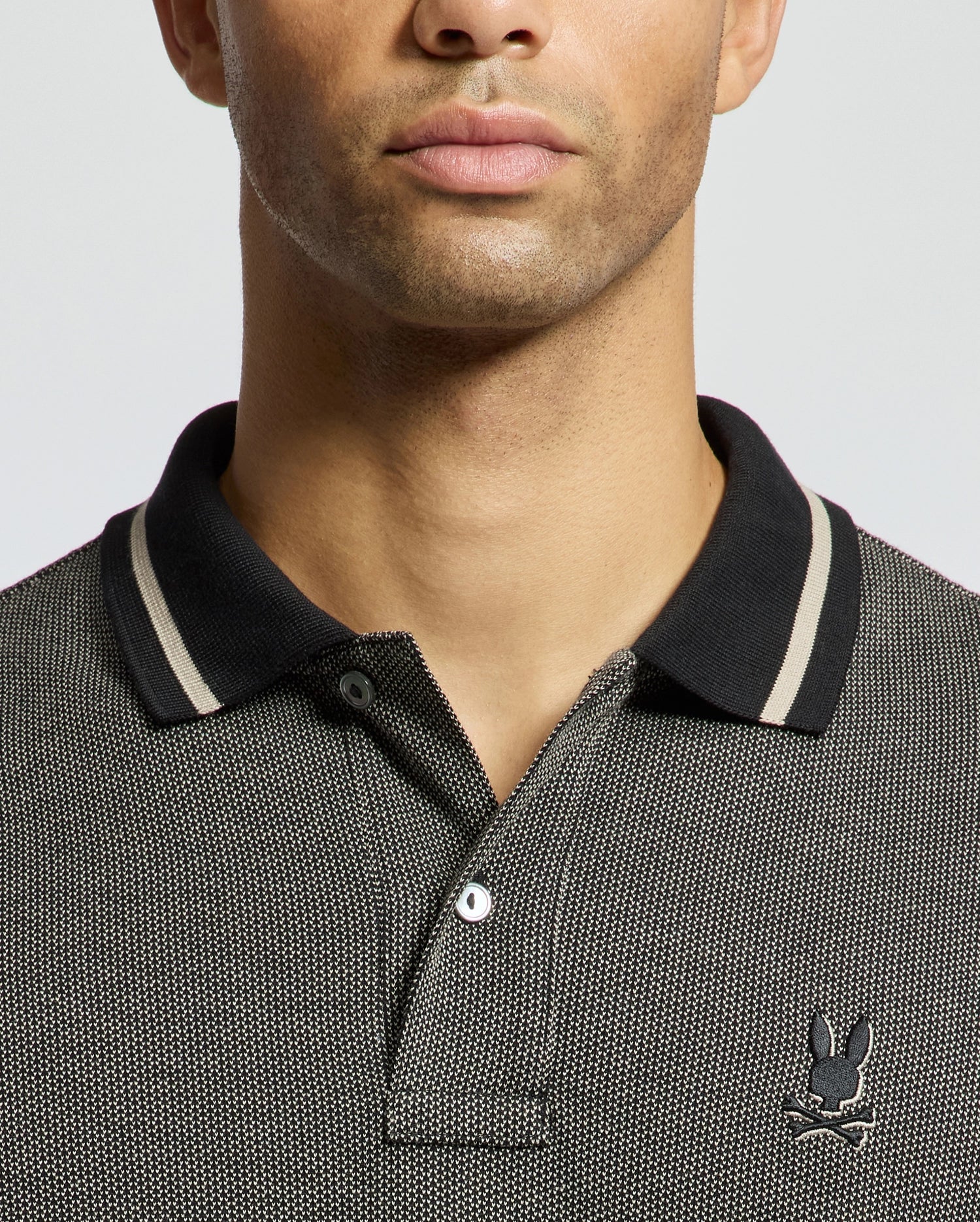 A close-up of a person wearing the Psycho Bunny "MENS WARSAW JACQUARD PIQUE POLO - B6K901E200" crafted from Peruvian Pima cotton, featuring a black and grey jacquard-knit design with a subtle checkered pattern. The polo shirt is accented with a black collar adorned with white stripes, and it has the brand's distinctive small embroidered logo of a rabbit with crossed bones. The person's face is partially visible in the image.