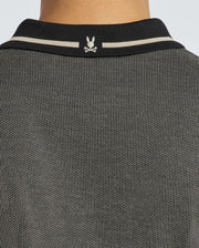 Close-up of a person's back wearing the Psycho Bunny Men's Warsaw Jacquard Pique Polo, featuring a textured, two-tone design crafted from jacquard-knit fabric with a black collar showcasing a small white logo. The collar includes two white stripes, and the fabric is woven with black and white threads.