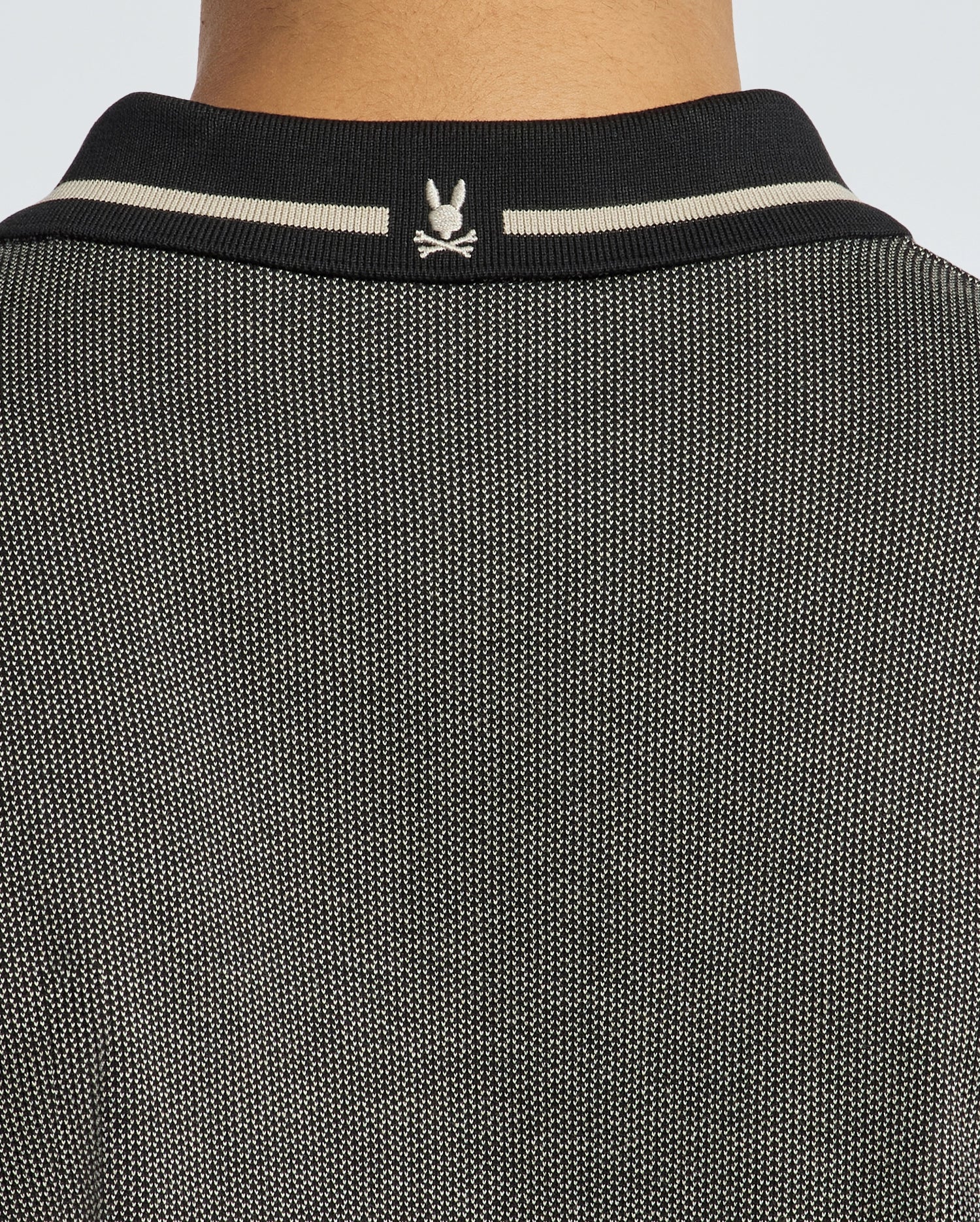 Close-up of a person's back wearing the Psycho Bunny Men's Warsaw Jacquard Pique Polo, featuring a textured, two-tone design crafted from jacquard-knit fabric with a black collar showcasing a small white logo. The collar includes two white stripes, and the fabric is woven with black and white threads.