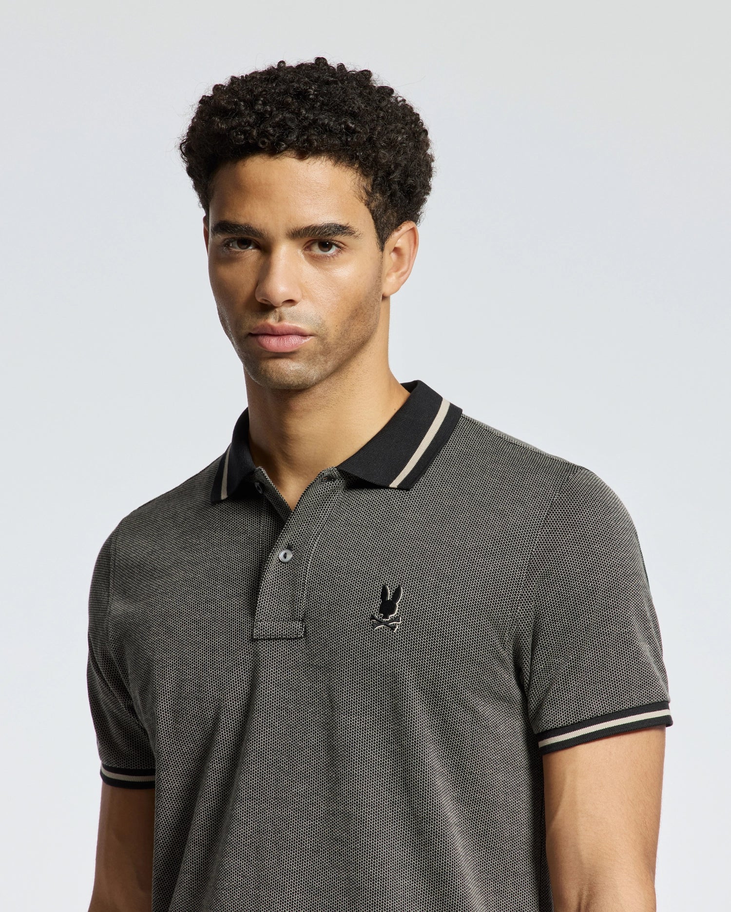 A man with short curly hair is wearing a gray, textured Psycho Bunny Men's Warsaw Jacquard Pique Polo, designed from luxurious Peruvian Pima cotton. It features black trim and a small emblem on the chest, set against a plain, light-colored background.