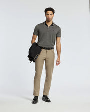 A man stands against a neutral background wearing a Psycho Bunny MENS WARSAW JACQUARD PIQUE POLO crafted from Peruvian Pima cotton paired with beige pants. He holds a dark jacket in his left hand and wears black loafers.