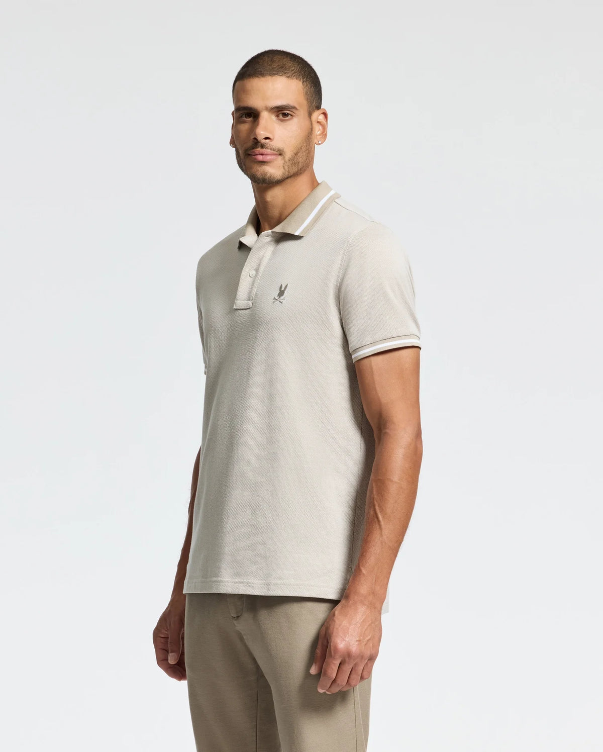 A man dressed in a light gray Psycho Bunny Men's Warsaw Jacquard Pique Polo, made from luxurious Peruvian Pima cotton and featuring an embroidered logo on the chest, stands against a plain white background. He has short dark hair and light stubble, with his hands by his sides, looking confidently at the camera.