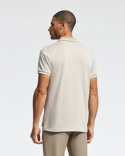 A man is seen from behind wearing a Psycho Bunny MENS WARSAW JACQUARD PIQUE POLO in light beige, crafted from luxurious Peruvian Pima cotton. The polo showcases a subtle collar design and pairs seamlessly with his pants against the plain white backdrop.