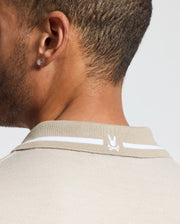 Close-up of a person wearing the Psycho Bunny Men's Warsaw Jacquard Pique Polo - B6K901E200, crafted from Peruvian Pima cotton in beige. It features a small embroidered rabbit head on the back. The individual has short hair and wears a stud earring in the visible ear.