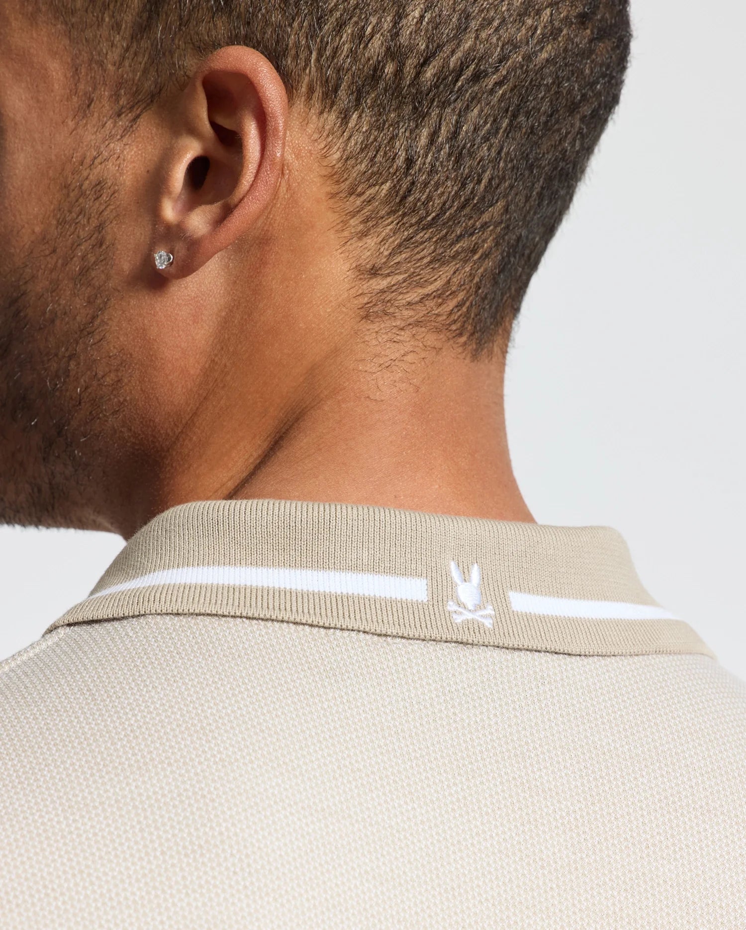 Close-up of a person wearing the Psycho Bunny Men's Warsaw Jacquard Pique Polo - B6K901E200, crafted from Peruvian Pima cotton in beige. It features a small embroidered rabbit head on the back. The individual has short hair and wears a stud earring in the visible ear.