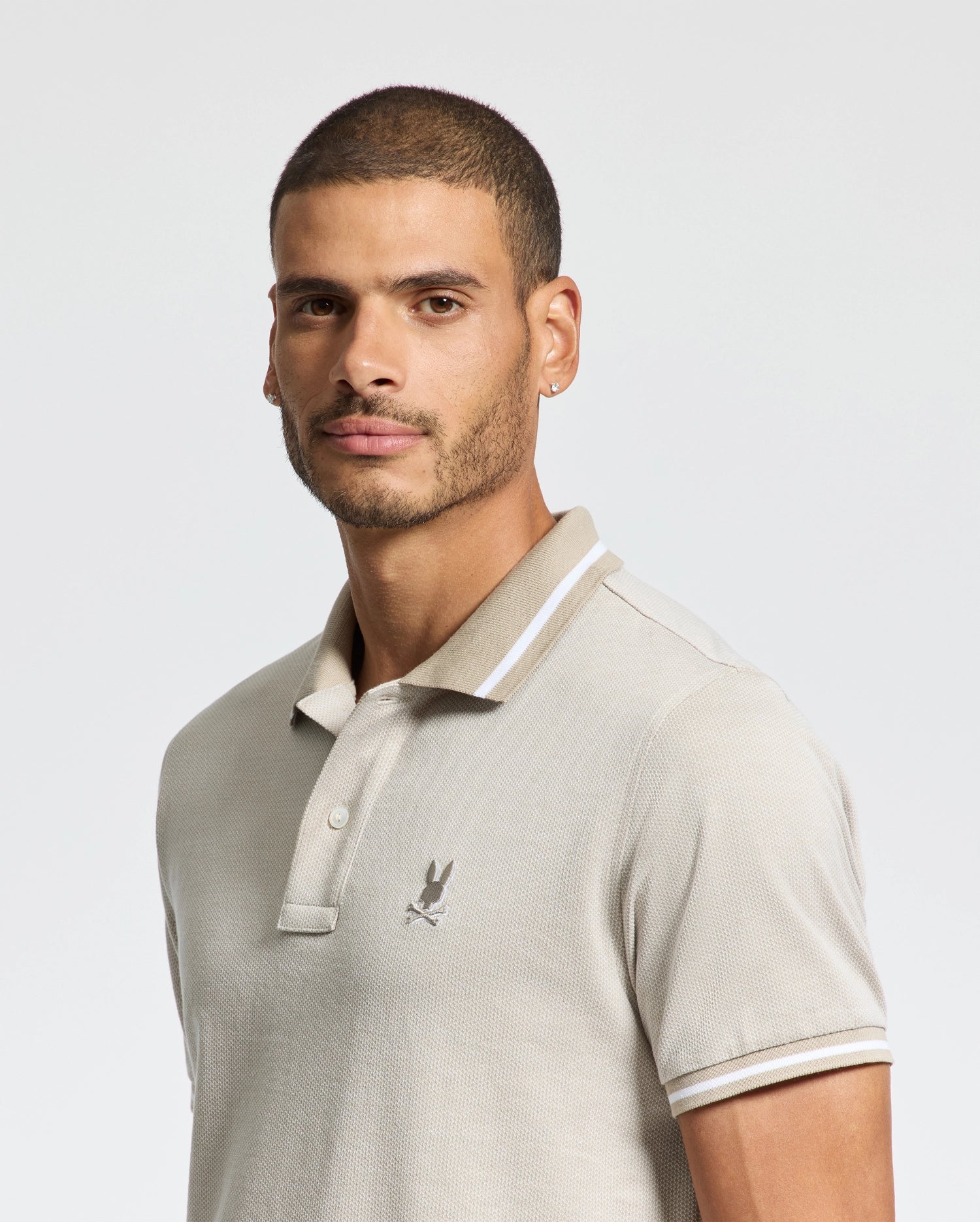 A person with short hair and a beard is wearing the Psycho Bunny MENS WARSAW JACQUARD PIQUE POLO - B6K901E200 in light gray, made from luxurious Peruvian Pima cotton. It features a small embroidered logo on the chest. The plain, light-colored background accentuates the shirt's classic collar and button-up neckline.