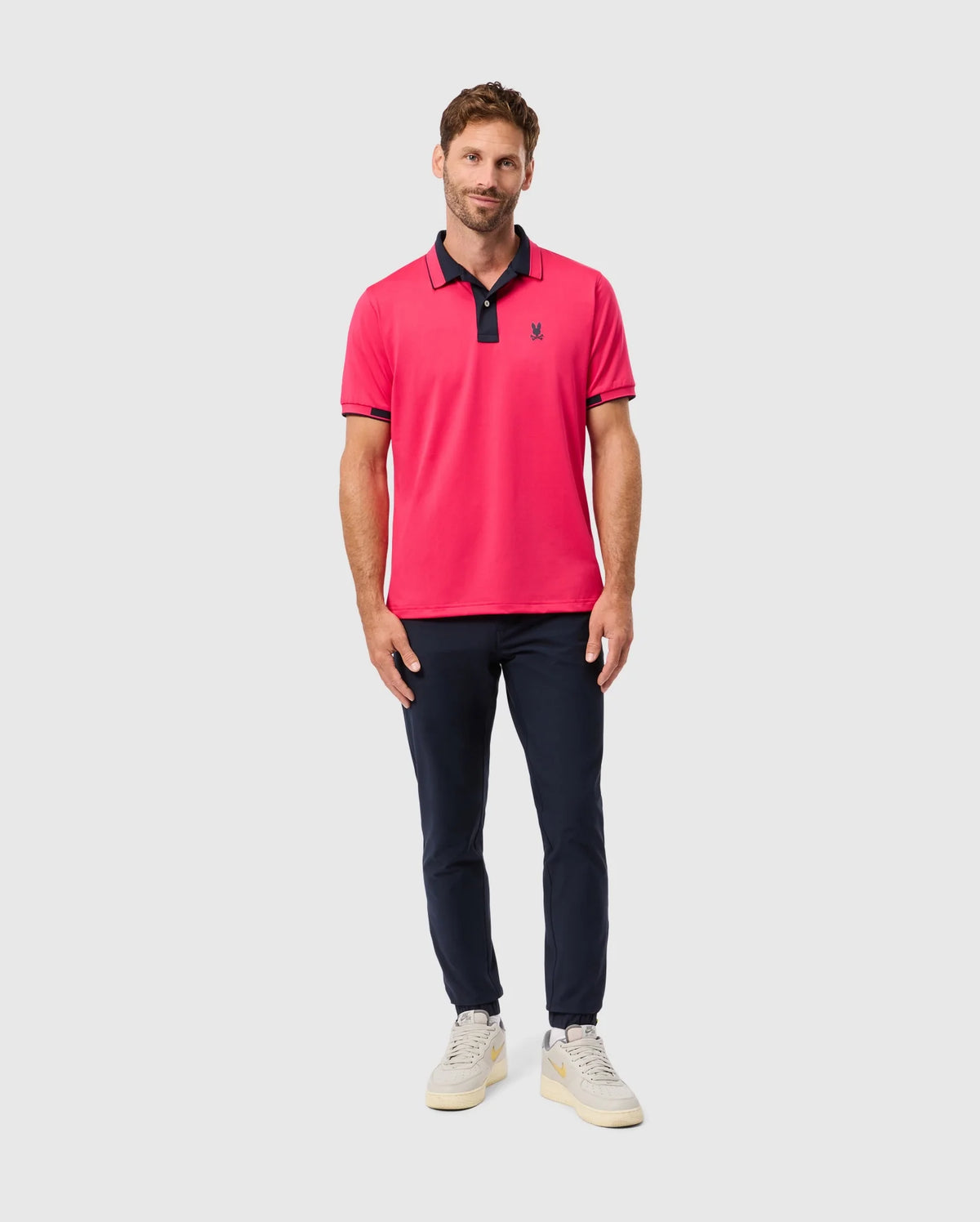A man is standing against a light gray background, wearing the Psycho Bunny MENS BELLEVUE SPORT POLO SHIRT - B6K905D200, featuring a bright pink color with dark trim. He pairs it with navy pants and white sneakers. With short brown hair and a light beard, he smiles gently.