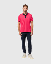 A man is standing against a light gray background, wearing the Psycho Bunny MENS BELLEVUE SPORT POLO SHIRT - B6K905D200, featuring a bright pink color with dark trim. He pairs it with navy pants and white sneakers. With short brown hair and a light beard, he smiles gently.