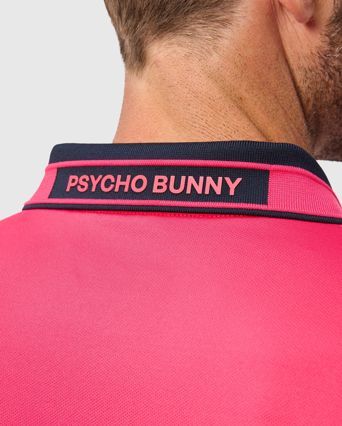 A close-up of a person wearing the MENS BELLEVUE SPORT POLO SHIRT - B6K905D200 from Psycho Bunny, showcasing its pink athletic knit with a contrasting black collar. The collar is embroidered with "PSYCHO BUNNY" in pink. The person's face is not visible, highlighting the shirt's breathable performance design and intricate detailing on the back and neck area.