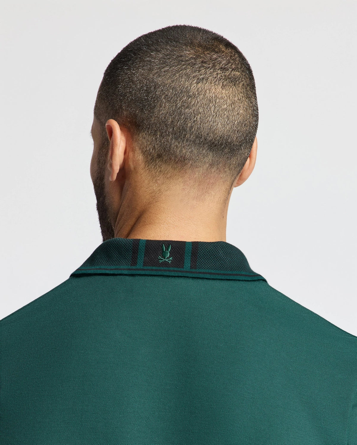 A person with a close-cropped haircut is facing away from the camera, wearing a Mens Malton Pique Polo from Psycho Bunny in dark green. Crafted from lightweight pique fabric, this high-quality men's polo features a small embroidered logo on the back of the collar. The background is light gray.