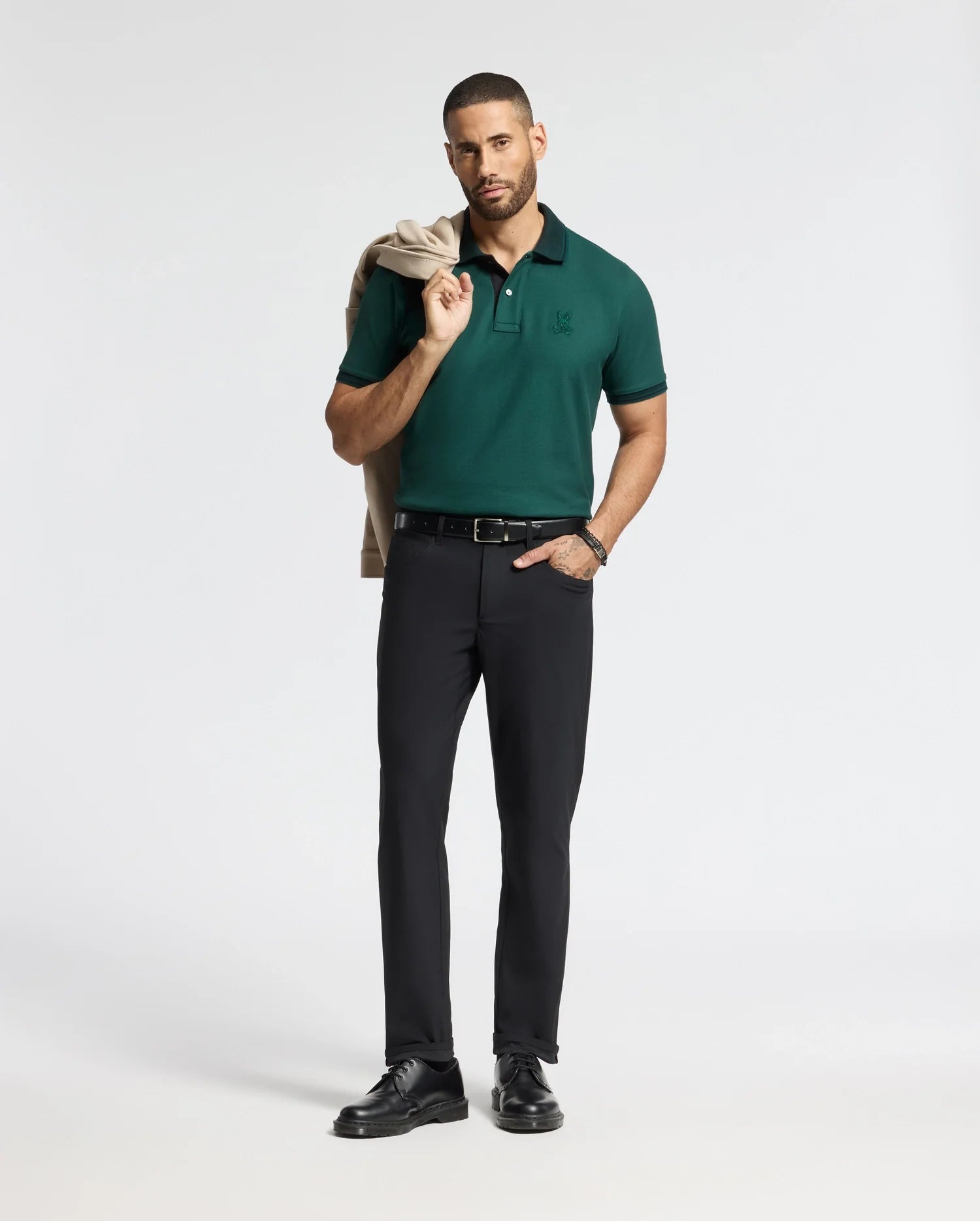 A man with a beard stands confidently, wearing the high-quality MENS MALTON PIQUE POLO by Psycho Bunny in green and black pants. He holds a folded beige jacket over his left shoulder and wears black shoes and a belt, set against a plain white background.