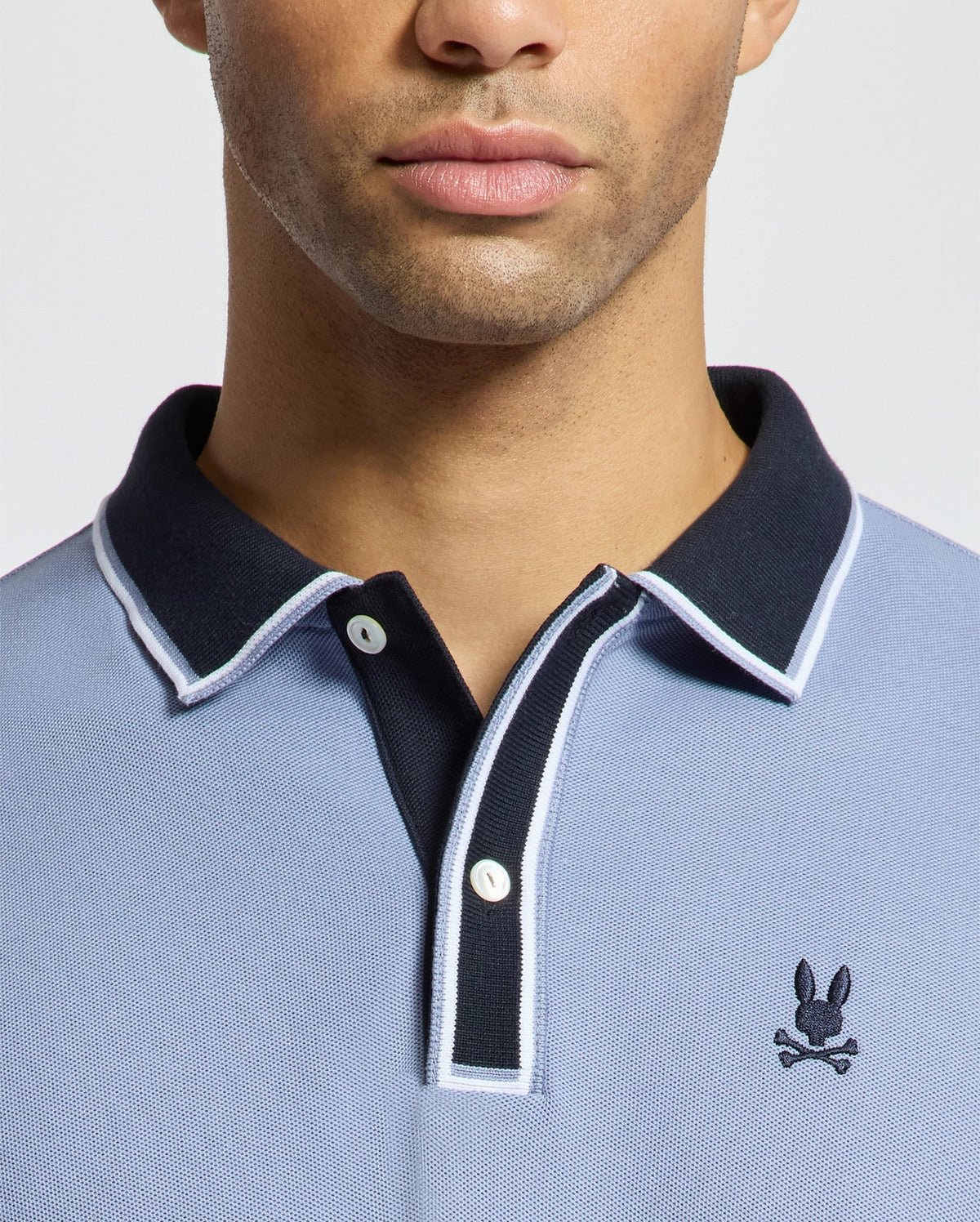 A person is wearing a light blue MENS KENT PIQUE POLO - B6K951D200 by Psycho Bunny, featuring a black collar and white trim. Made from premium materials, the shirt showcases a small emblem of a rabbit with crossed bones on the chest. The plain white background frames only the lower half of the face and upper torso.