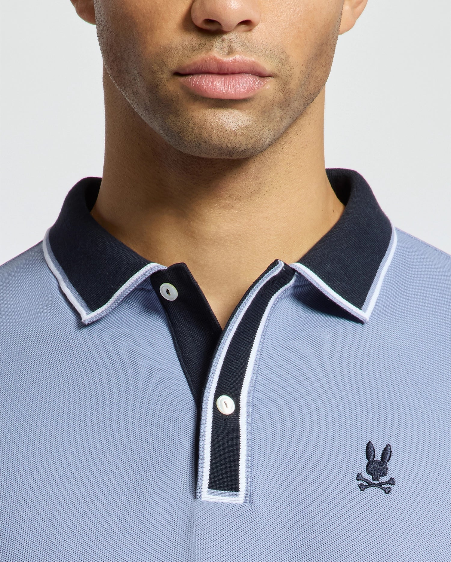A person is wearing a light blue MENS KENT PIQUE POLO - B6K951D200 by Psycho Bunny, featuring a black collar and white trim. Made from premium materials, the shirt showcases a small emblem of a rabbit with crossed bones on the chest. The plain white background frames only the lower half of the face and upper torso.