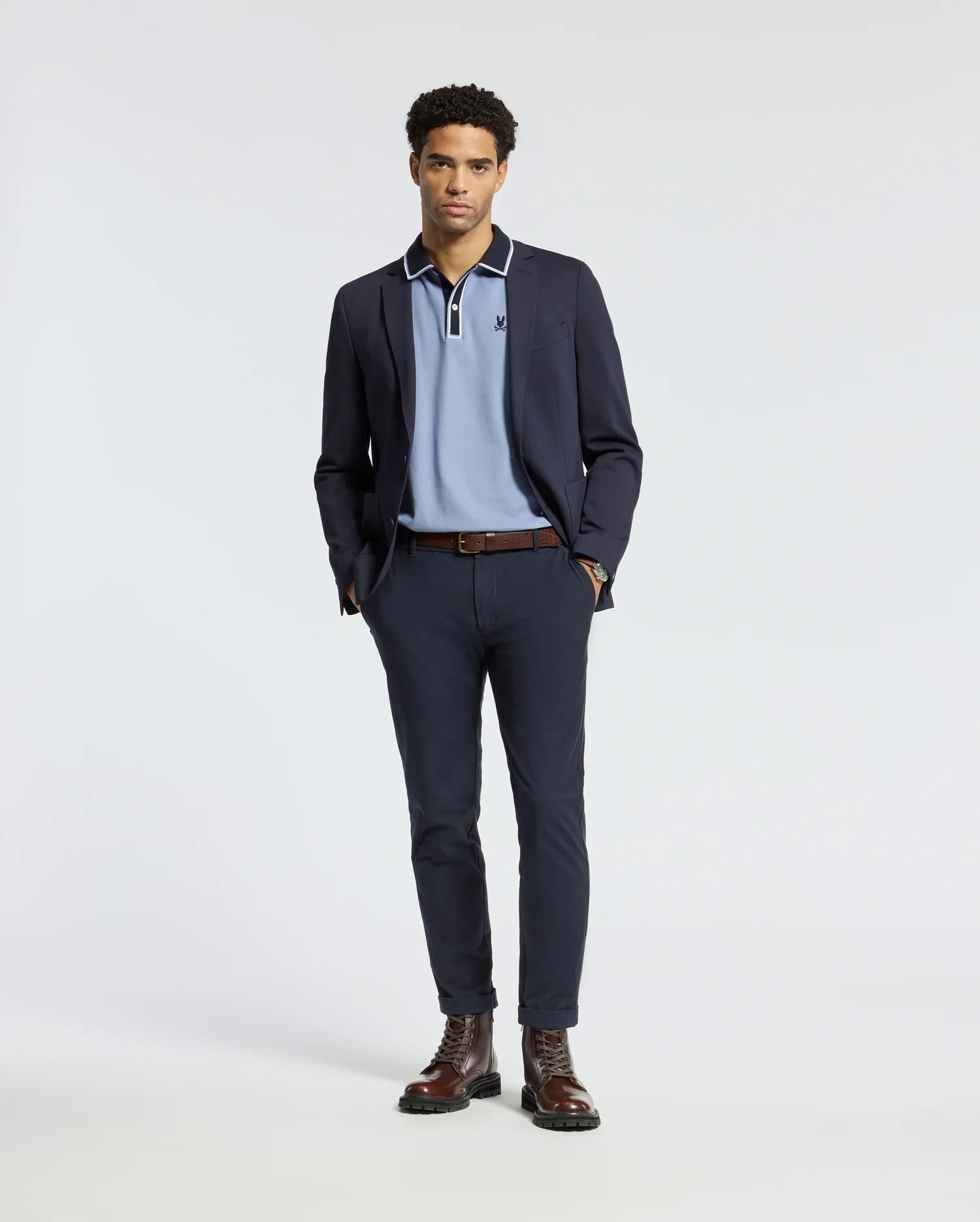 A man stands wearing a navy blazer, the Psycho Bunny MENS KENT PIQUE POLO - B6K951D200 in light blue, dark trousers, and brown boots. He has his hands in his pockets and poses against a plain white background.