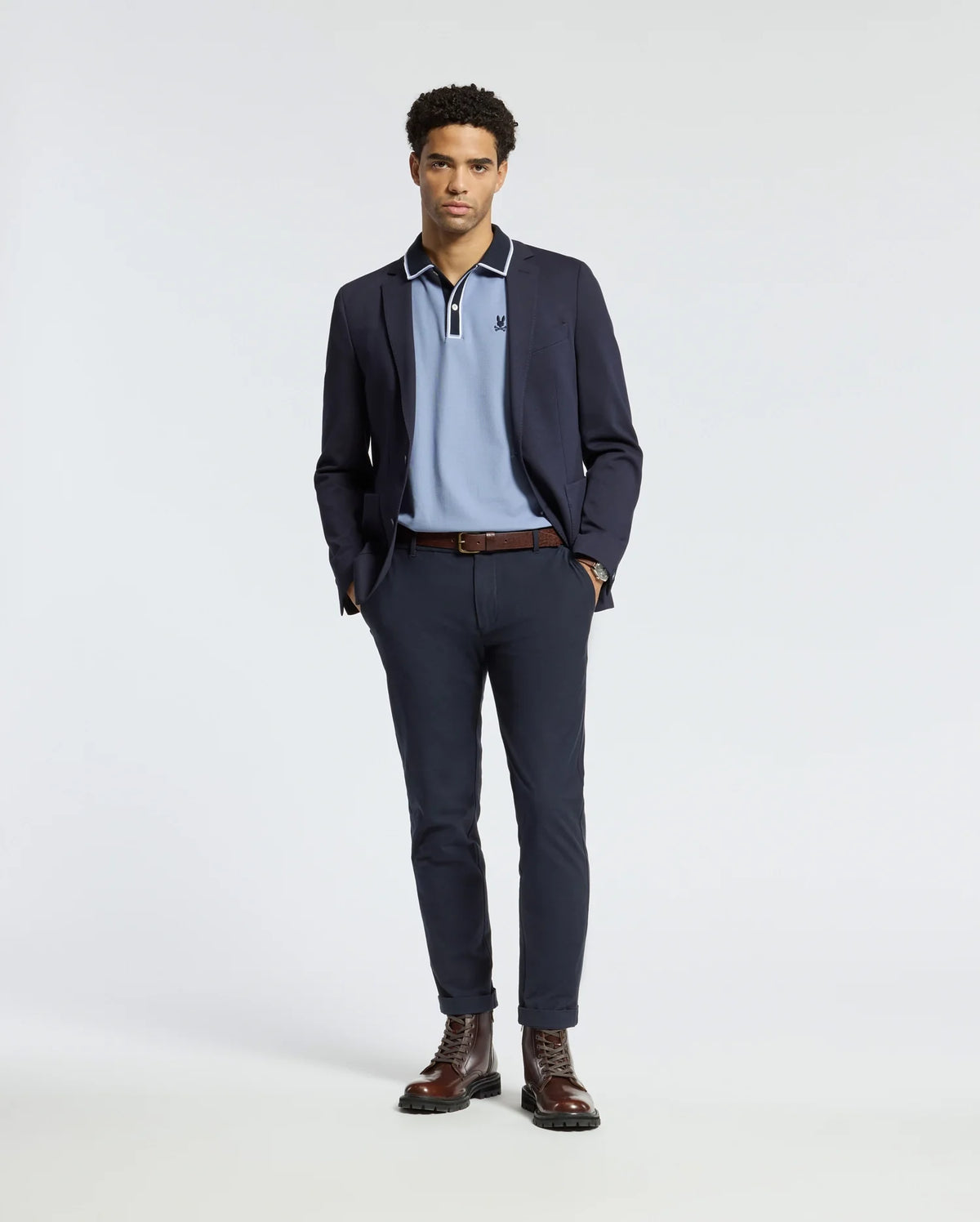 A man stands wearing a navy blazer, the Psycho Bunny MENS KENT PIQUE POLO - B6K951D200 in light blue, dark trousers, and brown boots. He has his hands in his pockets and poses against a plain white background.