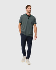A man stands casually, wearing the MENS KENT PIQUE POLO SHIRT by Psycho Bunny in dark green, featuring a small emblem. He pairs it with navy blue pants and white sneakers while his left hand rests in his pocket against a plain gray background.