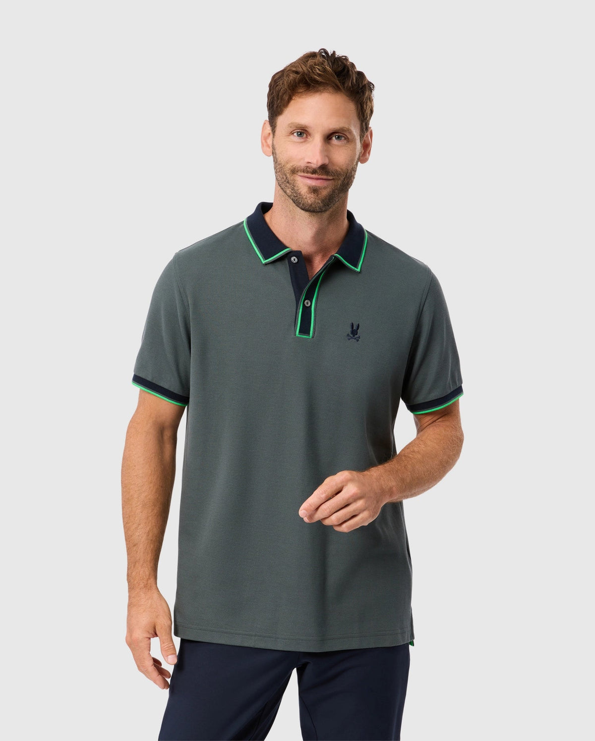 A man with short hair and a beard is wearing a stylish MENS KENT PIQUE POLO SHIRT - B6K951D200 by Psycho Bunny. The shirt features a chic dark green color with a black and green collar, made from premium Pima cotton. He pairs it with dark trousers and stands against a light gray background, looking forward with a slight smile while holding his fingers together.