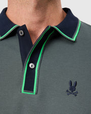 Close-up of a person wearing the MENS KENT PIQUE POLO SHIRT - B6K951D200 from Psycho Bunny, showcasing its stylish Pima cotton fabric in a gray hue. The polo features a dark navy collar and green trim, with an embroidered logo of a stylized skull and rabbit ears on the chest and two buttons at the collar.