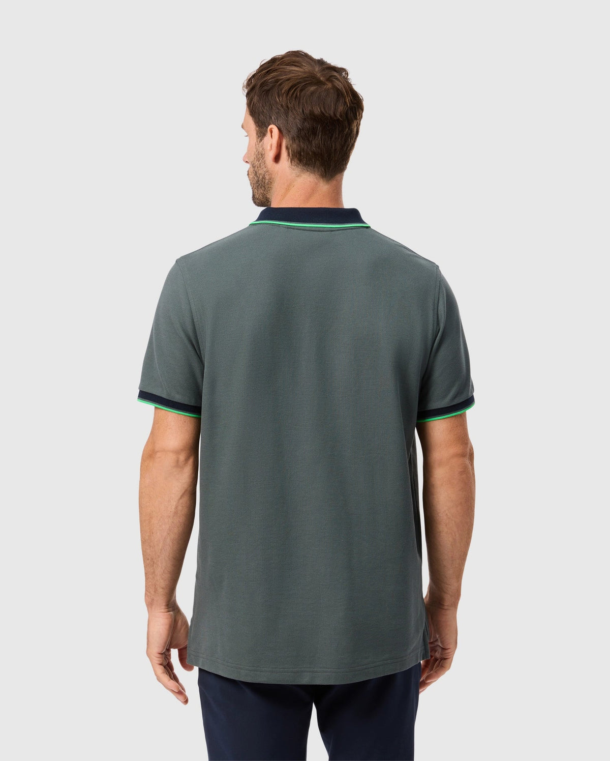 A man with brown hair is wearing a stylish Psycho Bunny polo in green, accented with black and green trim on the sleeves and collar. The MENS KENT PIQUE POLO SHIRT (B6K951D200), made from exquisite Pima cotton, exudes elegance as he stands facing away from the camera against a plain background.