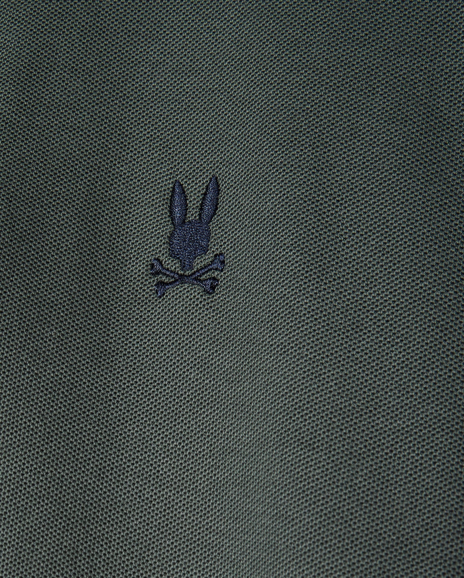 A close-up image of the textured dark green fabric from the Men's Kent Pique Polo Shirt by Psycho Bunny showcases a small embroidered logo, featuring a creative twist on the classic skull and crossbones design with a black silhouette of a rabbit's head above crossed bones.