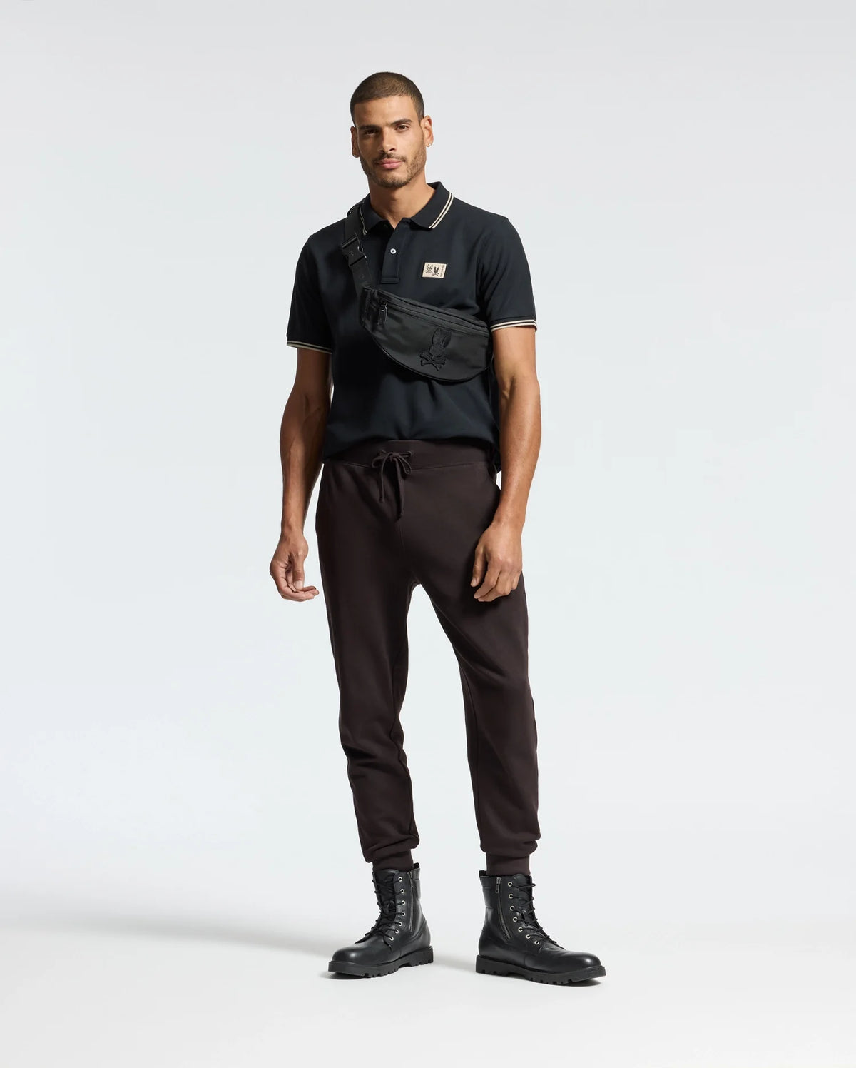A man stands against a simple backdrop wearing a black Monroe Pique Polo from Psycho Bunny, paired with dark brown jogger pants and black lace-up boots. Draped across his chest is a black crossbody bag featuring Mother-of-Pearl buttons. His expression is neutral, while his arms rest easily by his sides.