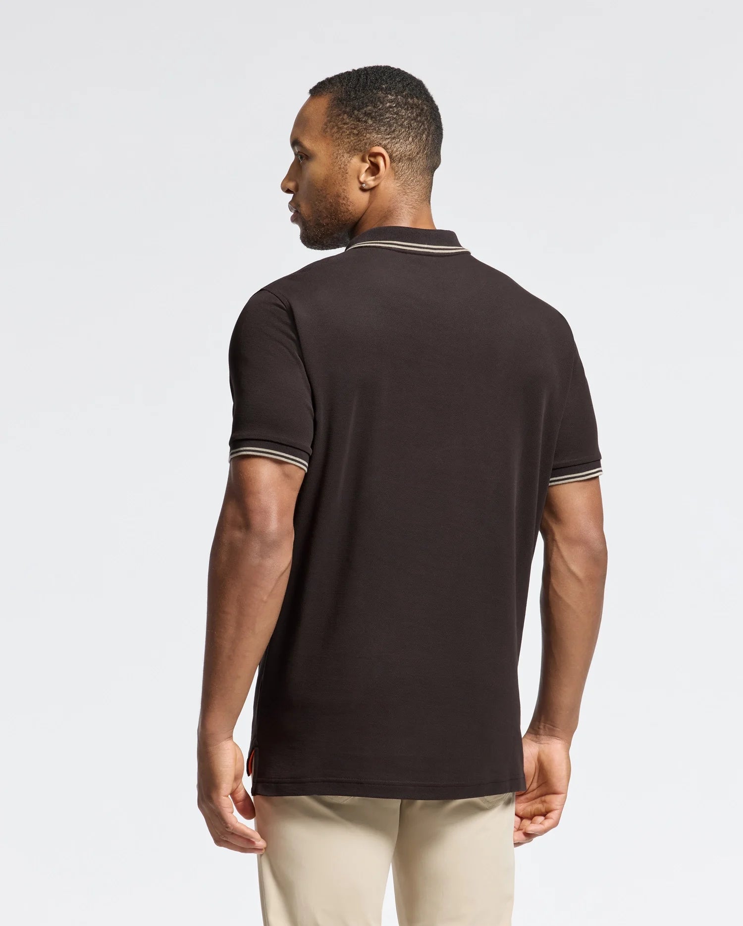A man is standing with his back to the camera, wearing a Psycho Bunny MENS MONROE PIQUE POLO - B6K964D200 in black, which features mother-of-pearl buttons, paired with beige pants. The background is a neutral light gray.