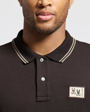 A person is wearing the Psycho Bunny MENS MONROE PIQUE POLO - B6K964D200, a dark Pima cotton polo in Big and Tall sizes, featuring double stripes on the collar and a patch with two cartoon skulls on the left side. The background color of the shirt is light gray.