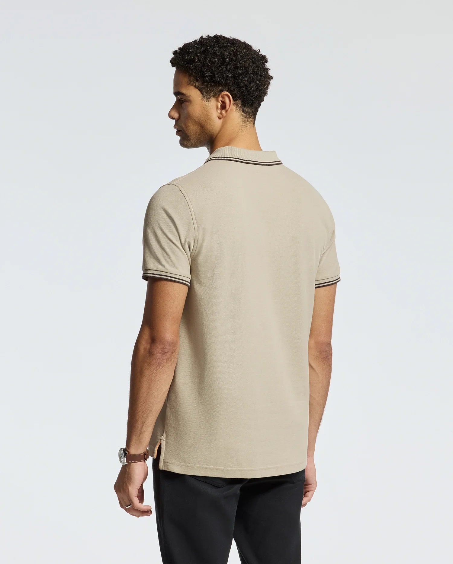 A man with curly hair, wearing the Psycho Bunny Men's Monroe Pique Polo in beige with dark trim and regular fit black pants, stands facing away while glancing to the side. The plain light gray background highlights the quality craftsmanship of this polo shirt, made in Peru.