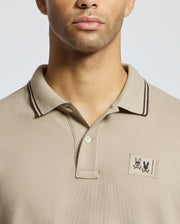 A person wearing a beige Monroe Pique Polo by Psycho Bunny, featuring dark trim on the collar. The shirt, crafted from Pima cotton in Peru, showcases a small floral patch and logo on the left side. The background is a neutral light gray.