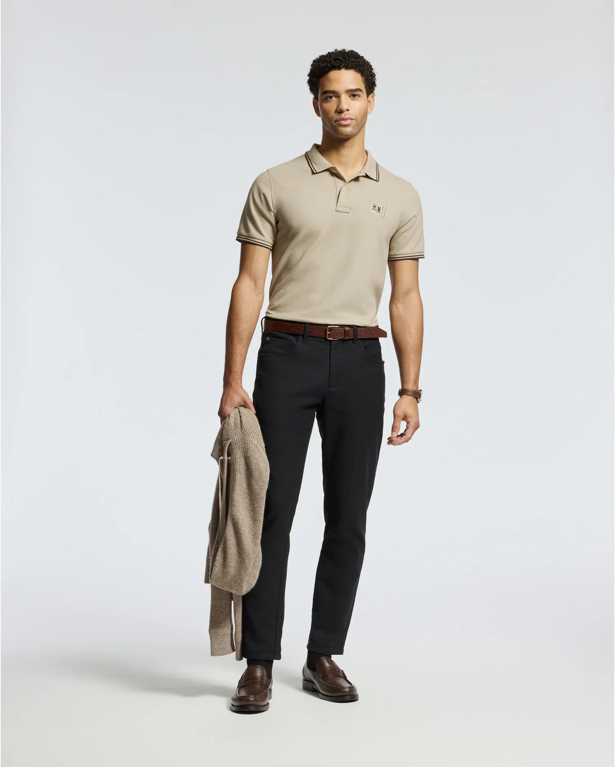 A man stands against a plain background, wearing a Psycho Bunny MENS MONROE PIQUE POLO in light beige, black pants, and brown shoes. Made in Peru, the regular fit polo exudes casual elegance. He holds a light brown jacket in his left hand, his expression neutral, and his short hair styled simply.