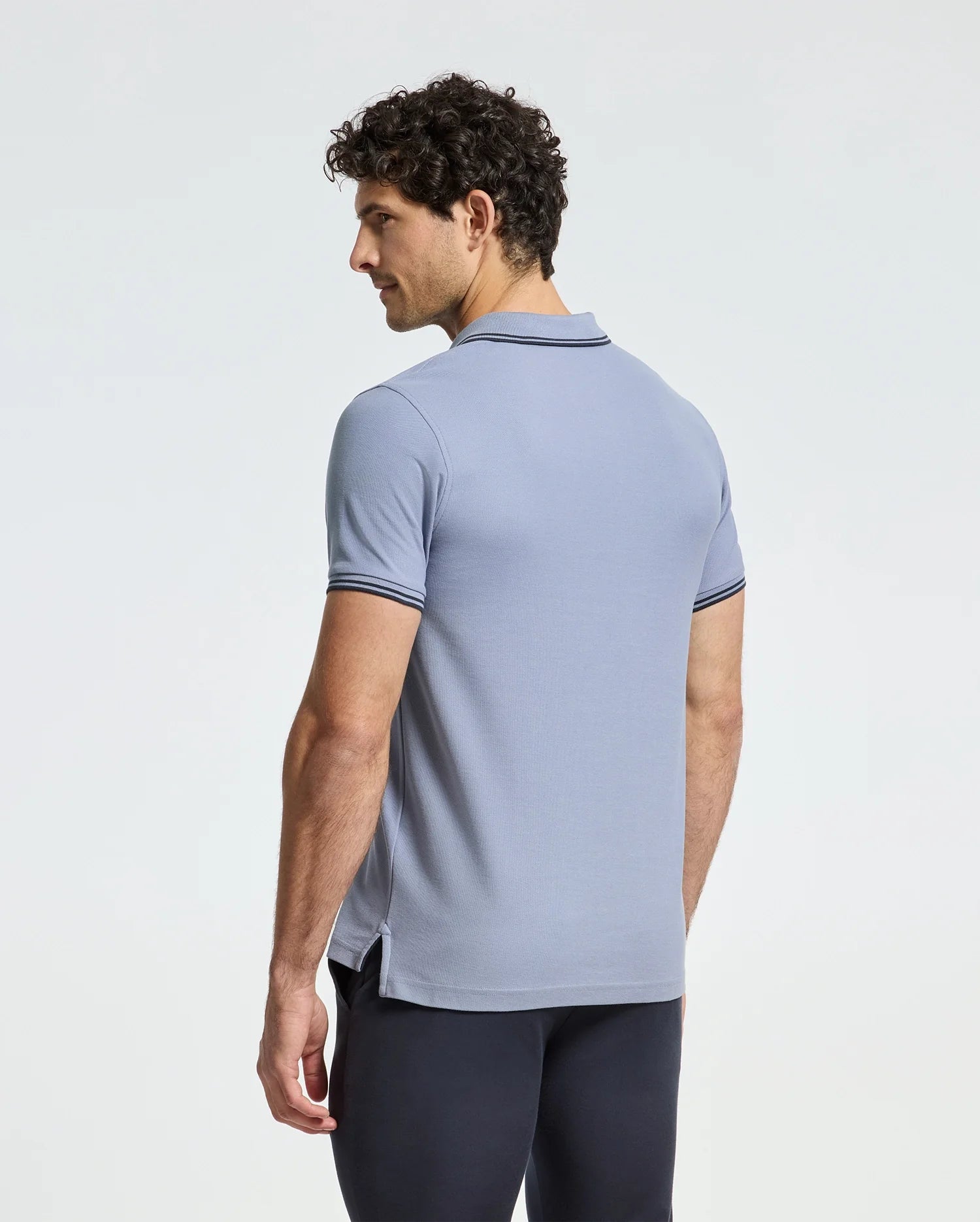 A man with curly hair is wearing a Psycho Bunny Monroe Pique Polo, featuring regular fit with Mother-of-Pearl buttons and dark trim, paired with black pants. He stands slightly turned to the side against a plain light background.