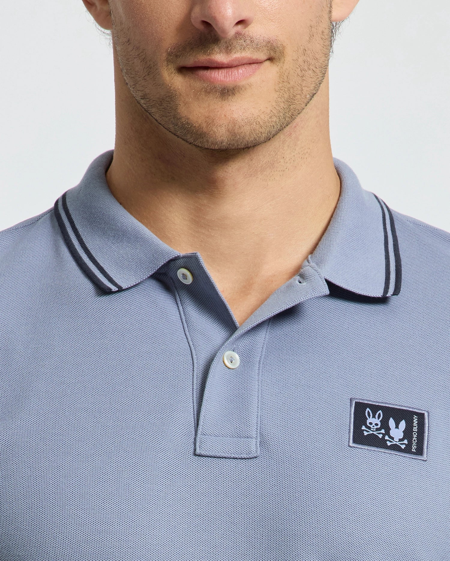A man models the Psycho Bunny MENS MONROE PIQUE POLO - B6K964D200, made from soft light blue Pima cotton. It features an embroidered skull and crossbones on the chest, Mother-of-Pearl buttons, and a dark-trimmed collar, showcasing his upper torso and part of his face.