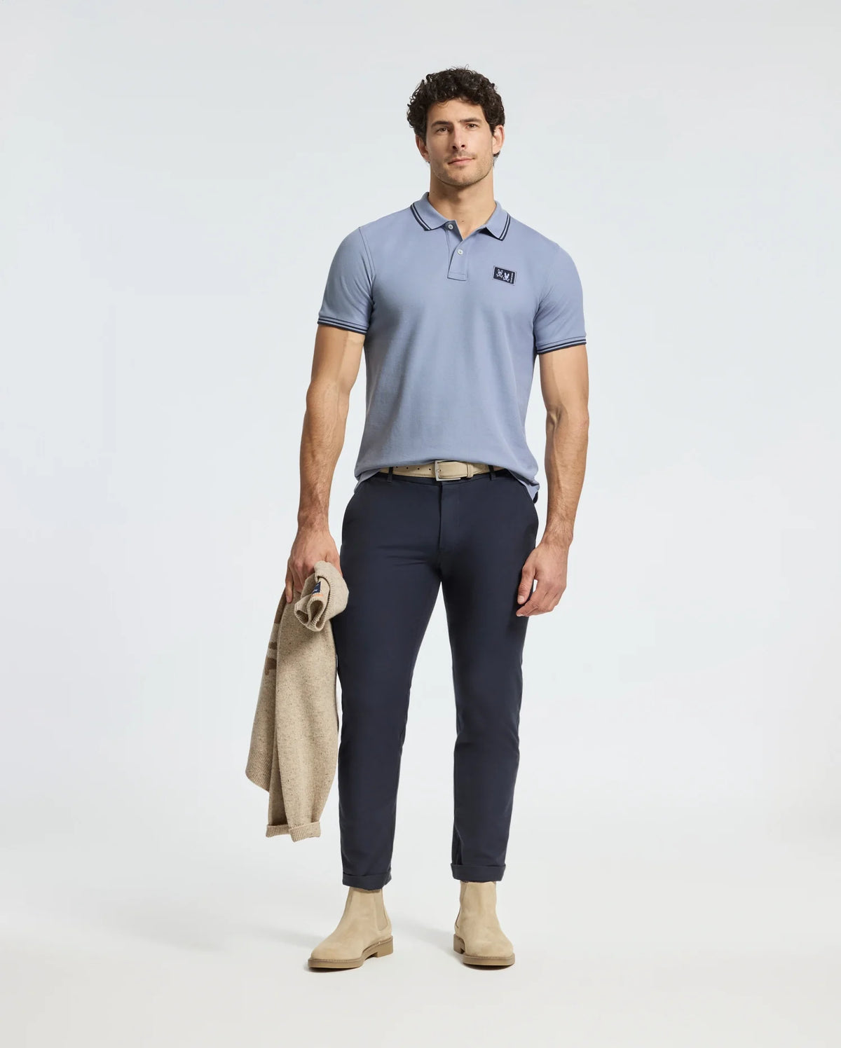 A man in a Psycho Bunny MENS MONROE PIQUE POLO with Mother-of-Pearl buttons stands holding a beige jacket. He wears dark blue pants, a beige belt, and matching shoes, posing against a light background.