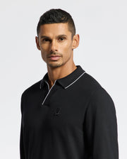 A person with short hair is wearing a black Psycho Bunny MENS CREEDE PIQUE JOHNNY COLLAR POLO - B6M265E200, which features a small embroidered logo on the chest. The Pima cotton shirt's collar has white piping, creating a subtle contrast against the plain, light-colored background.