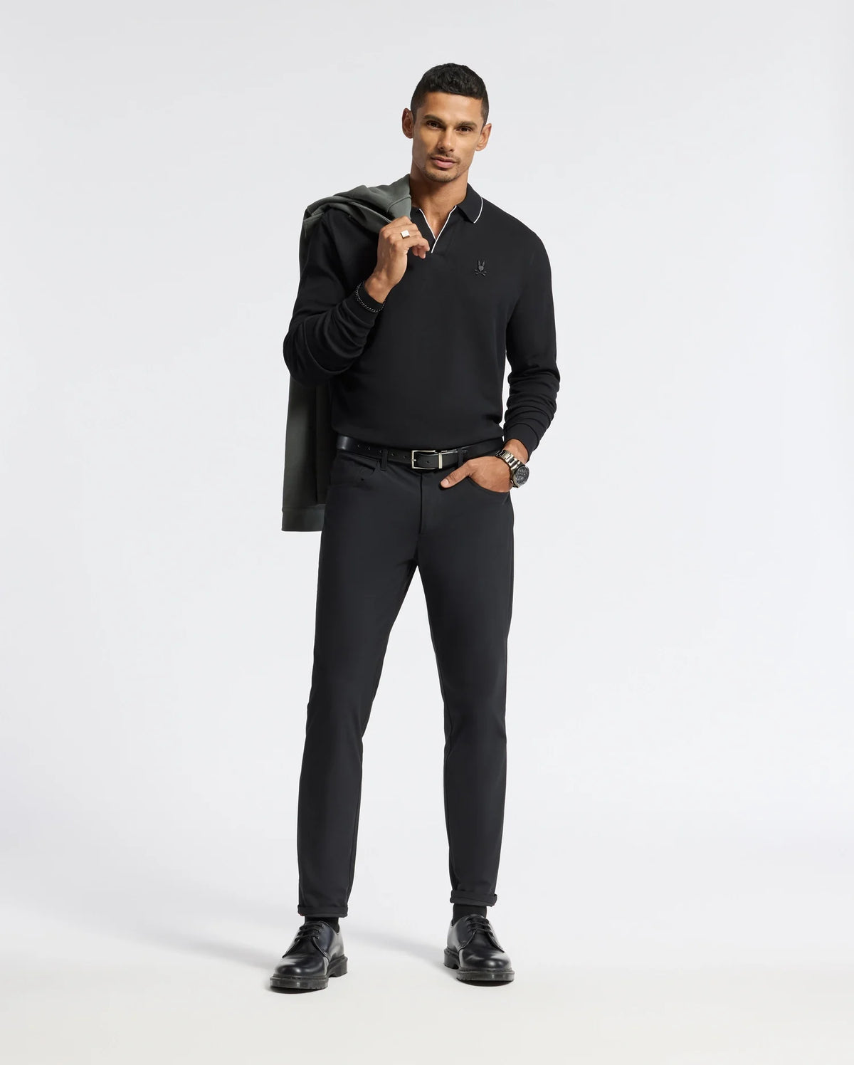 A man in a sleek black outfit poses confidently, showcasing a Psycho Bunny MENS CREEDE PIQUE JOHNNY COLLAR POLO alongside tailored pants and shoes. Adding a stylish touch, he casually drapes a jacket over his shoulder against a plain white background.