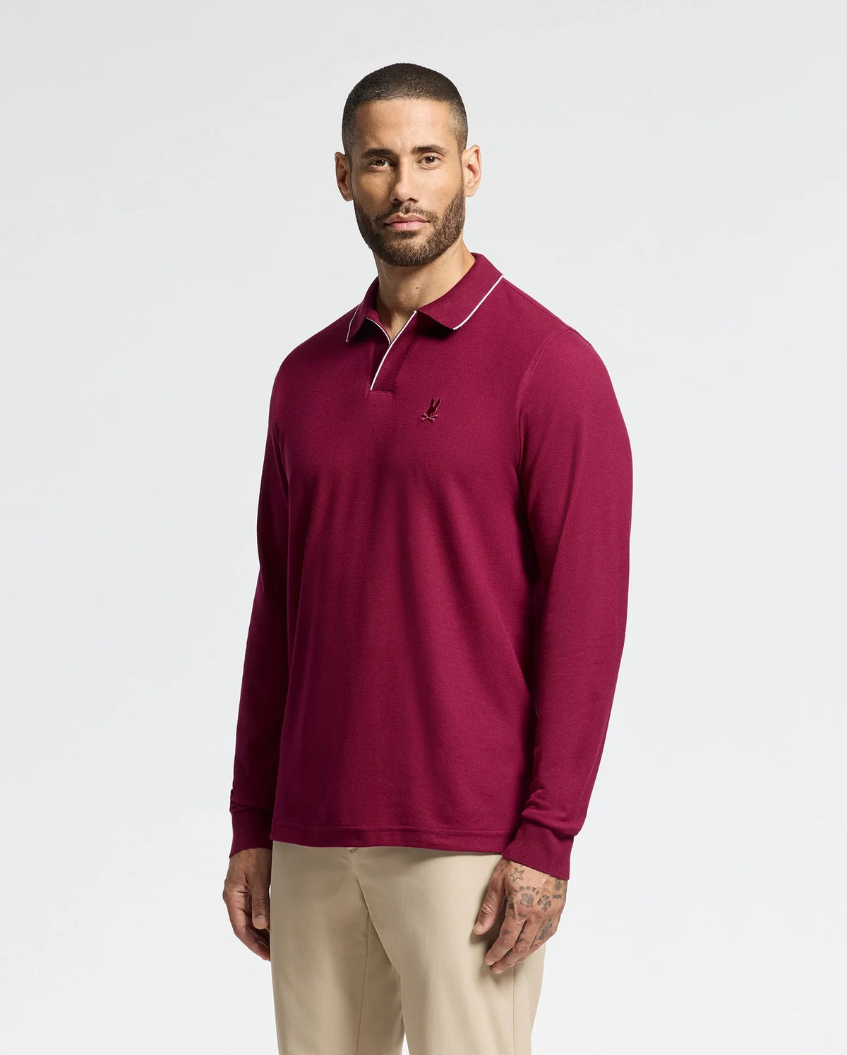 A person wearing a burgundy Psycho Bunny MENS CREEDE PIQUE JOHNNY COLLAR POLO - B6M265E200 with a small embroidered logo on the chest stands against a plain white background. They have short hair and a beard, and are also wearing beige pants.