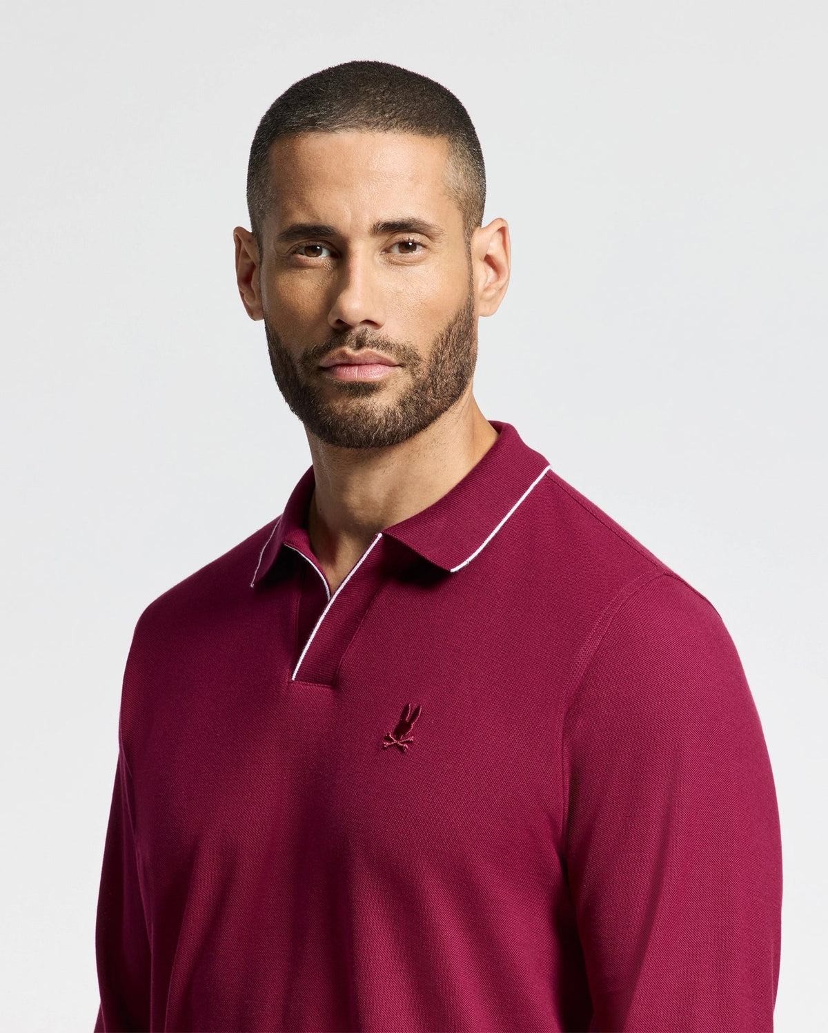 A man with short hair and a beard is wearing the Psycho Bunny Men's Creede Pique Johnny Collar Polo in burgundy, featuring white trim and a small embroidered logo on the chest. He is looking slightly to the side against a plain, light background.