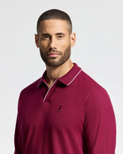 A man with short hair and a beard is wearing the Psycho Bunny Men's Creede Pique Johnny Collar Polo in burgundy, featuring white trim and a small embroidered logo on the chest. He is looking slightly to the side against a plain, light background.