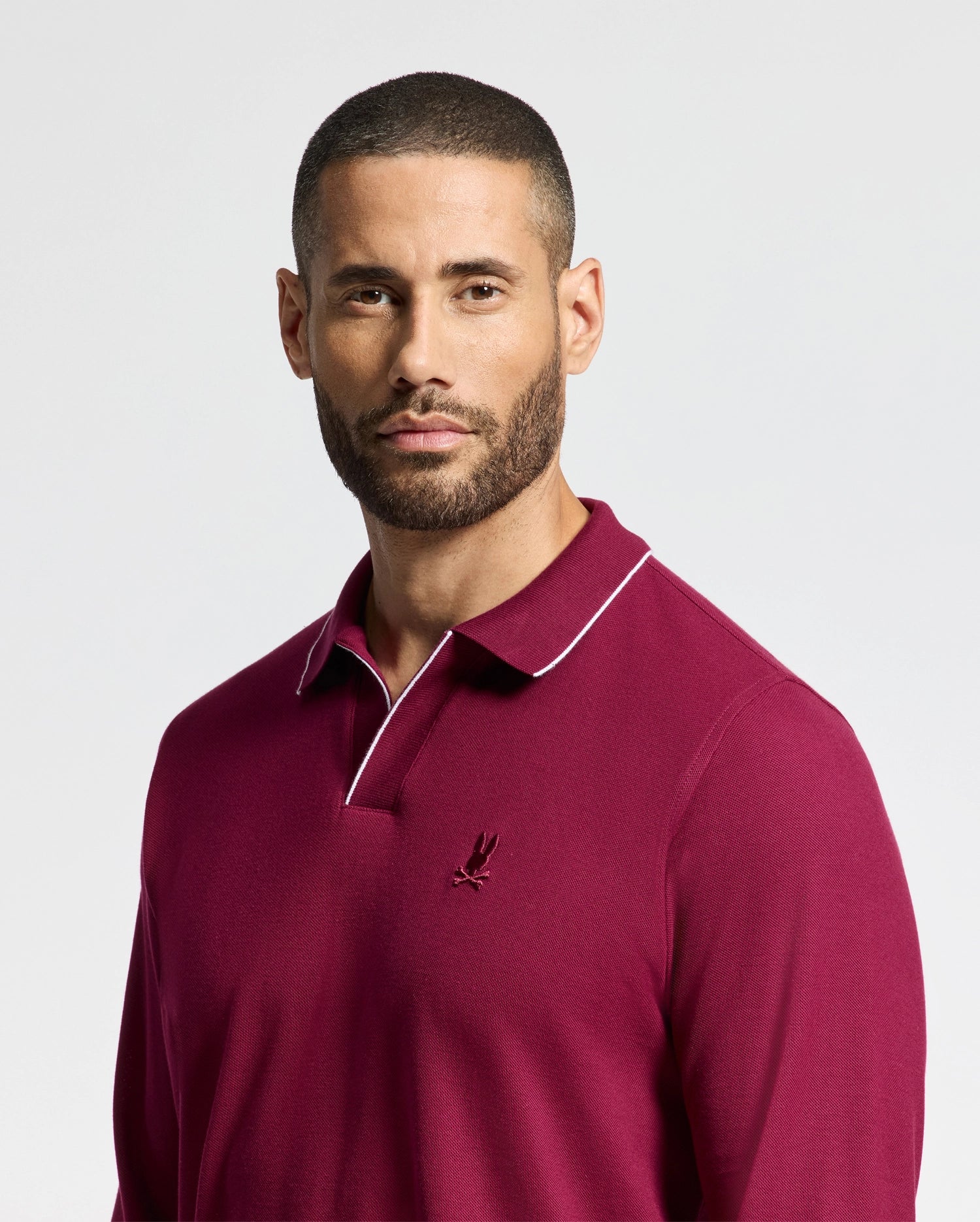 A man with short hair and a beard is wearing the Psycho Bunny Men's Creede Pique Johnny Collar Polo in burgundy, featuring white trim and a small embroidered logo on the chest. He is looking slightly to the side against a plain, light background.