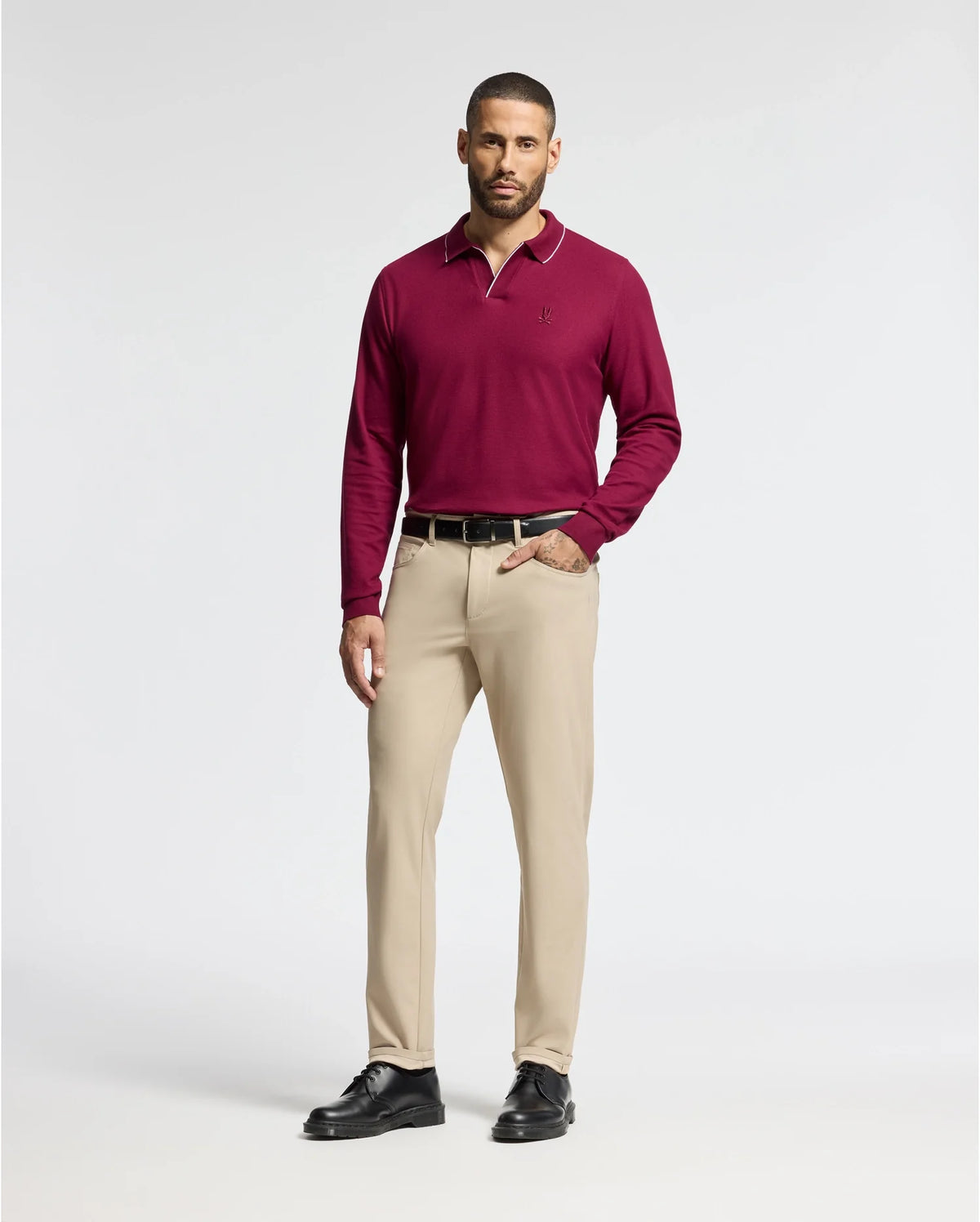 A man with short hair, dressed in a burgundy Psycho Bunny MENS CREEDE PIQUE JOHNNY COLLAR POLO - B6M265E200 and beige trousers, stands with one hand in his pocket against a plain background. The Pima cotton polo enhances the look with sophistication, perfectly complemented by his black shoes.