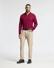 A man with short hair, dressed in a burgundy Psycho Bunny MENS CREEDE PIQUE JOHNNY COLLAR POLO - B6M265E200 and beige trousers, stands with one hand in his pocket against a plain background. The Pima cotton polo enhances the look with sophistication, perfectly complemented by his black shoes.